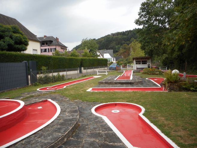 Minigolfanlage in Vianden mit roten Bahnen, Steinwegen, Bäumen und Häusern im Hintergrund.