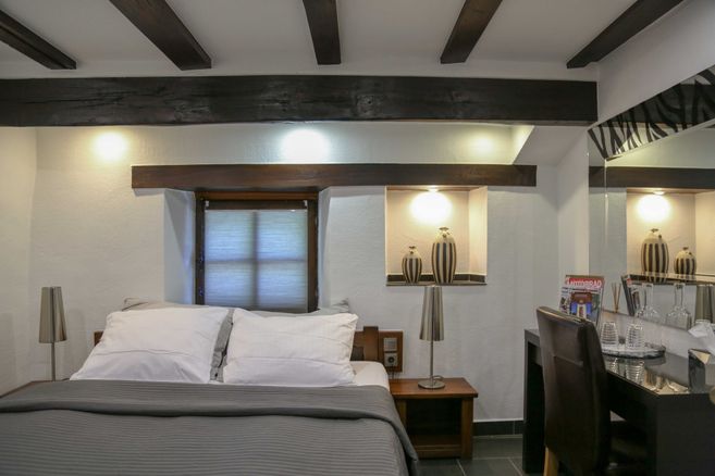 Ein gemütliches Hotelzimmer mit einem Doppelbett, Holzbalken an der Decke, einem kleinen Tisch mit Stuhl und dekorativen Vasen in einer Nische.